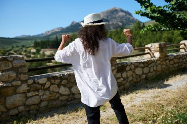 Kıvırcık saçlı, beyaz gömlekli ve şapkalı bir kadının arka planda dağlarla dolu güzel bir kırsalda eğlenişi..