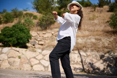 Doğada neşeli bir şekilde dans eden beyaz bir gömlek ve şapka giymiş, güneşin altında güneşlenen bir kadın..