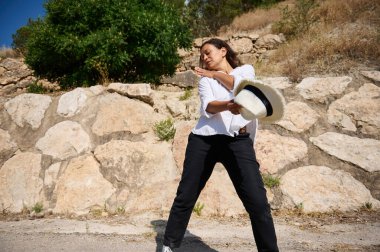 Güneşli bir günde açık havada beyaz gömlek ve siyah pantolon giyen bir kadın, kayalık bir duvarın ve yeşil bitkilerin yanında duruyor..