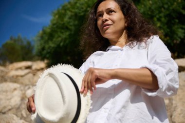 Güneşli bir günde dışarıda dikilirken beyaz gömlek giyen bir kadın şapkasını tutar. Yukarıdaki mavi gökyüzü ile birlikte doğal ortamda rahat ve mutlu görünüyor. Yaşam tarzı kavramları için ideal.