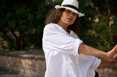 Parkta uzanan beyaz gömlekli ve şapkalı bir kadın. Güneş ışığı ve yeşil arka plan, dingin ve rahat atmosfere katkı sağlar..