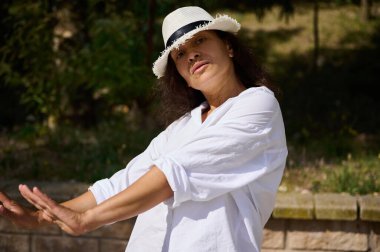 Beyaz gömlekli ve şapkalı bir kadın, güneşli bir günde doğal park ortamında eğleniyor. Rahatlamayı, kendine güveni ve günlük yaşam tarzını ifade eder.