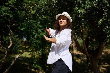 Güneşli bir günde yemyeşil bir parkta dans eden ve eğlenen mutlu bir kadın. Beyaz bir şapka ve gömlek giyiyor, neşe ve özgürlük saçıyor..