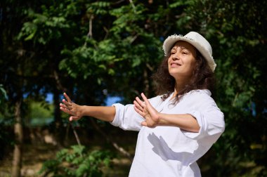 Parkta kollarını açmış, beyaz bir şapka ve gömlek giymiş özgürlüğünü ve mutluluğunu ifade eden doğanın tadını çıkaran bir kadın. Arka planda yemyeşil ağaçlar, yüzünü aydınlatan güneş ışığı..
