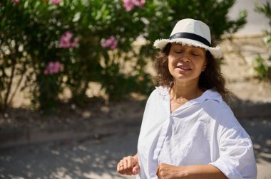 Gülümseyen kadın açık havada güneşli bir günün tadını çıkarıyor, beyaz bir gömlek ve şapka giyiyor, yeşil ve arka planda çiçekler var..