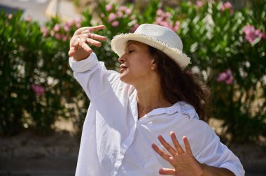 Beyaz gömlekli, şapkalı, gözleri kapalı bir kadın, açık havada güneşli bir günün tadını çıkarıyor. Arka planda yeşil çalılar ve pembe çiçekler var..