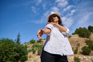 Kollarını açmış, açık havada güneşli bir günün tadını çıkaran beyaz bir gömlek ve şapka giyen, doğal bir manzaraya sahip bir kadın..