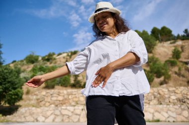 Beyaz gömlek giyen ve şapka takan bir kadın güneşli bir günde açık havada doğal bir zemine karşı tai chi antrenmanı yapıyor. Huzurlu ve rahat.