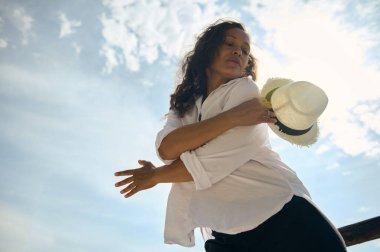 Beyaz gömlekli ve hasır şapkalı bir kadın güneşli bir açık havada rahatlıyor ve bulutlu mavi bir gökyüzünün altında kaygısız..