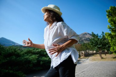 Dağlık bir bölgede beyaz gömlekli ve şapkalı bir kadın doğanın tadını çıkarıyor. Bir rahatlama anı yakalar ve doğayla bağlantı kurar..