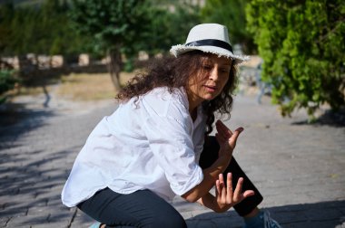 Açık hava parkında dans eden genç bir kadın, beyaz şapkalı ve günlük kıyafetli. Doğal çevre ve güneş ışığı canlı atmosferi güçlendirir..