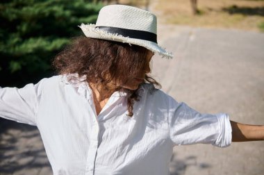 Beyaz gömlekli ve hasır şapkalı bir kadın açık havada güneşli bir günün tadını çıkarırken kollarını uzatıyor, özgürlük ve rahatlama taşıyor..