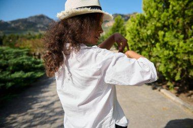 Beyaz gömlekli ve şapkalı bir kadın açık havada güneşli bir günün tadını çıkarıyor. Etrafı yemyeşil ve manzaralı dağ manzaralarıyla çevrili. Kameraya arkası dönük poz veriyor..