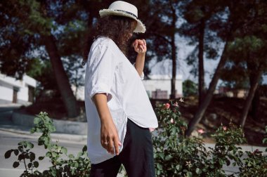 Beyaz gömlekli ve şapkalı bir kadın güneşli bir günde dışarıda yürüyor. Etrafı yeşillik ve ağaçlarla çevrili, doğanın tadını çıkarıyor..