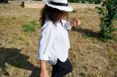 Beyaz gömlek ve hasır şapka giyen bir kadın güneşli bir günde çimenli bir tarlada yürüyor, rahat ve rahat bir yaşam tarzı sergiliyor..