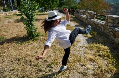 Güneşli bir günde, açık havada dans eden neşeli bir kadın, beyaz bir şapka ve günlük kıyafetler giyiyor. Arka planda yeşil ve taş çitli bir park var..