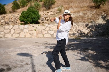 Kendine güveni tam bir kadın açık havada şık bir poz verir. Günlük kıyafetler ve şapka giyer. Güneşli bir ortamda taş bir duvar ve ağaçlarla birlikte..