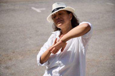 Beyaz şapkalı ve gömlekli bir kadın dışarıda dans ediyor, sıcak havanın tadını çıkarıyor. Güneşli bir günün neşe ve özgürlüğünü yakalar.