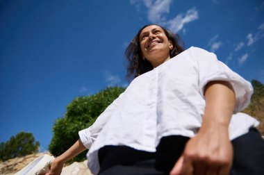 Neşeli bir kadın açık havada güneşli bir günün tadını çıkarırken ışıl ışıl gülümser. Düşük bir açıdan, açık mavi bir gökyüzünün arka planına ve yemyeşil bir alana doğru çekilmiş..