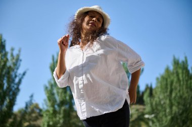 Beyaz gömlek ve şapkayla güneşli bir günün tadını çıkaran bir kadın. Arka planda parlak mavi bir gökyüzü ve yemyeşil, neşe ve özgürlük aktarılıyor..