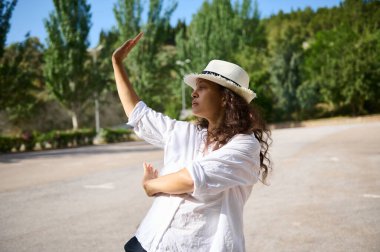 Bereli ve beyaz tişörtlü bir kadın, yemyeşil ve açık mavi gökyüzü olan huzurlu bir parkta tai chi çalışıyor..
