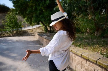 Güneşli bir günde, açık havada poz veren beyaz gömlekli ve şapkalı bir kadın. Arka planda ağaçlar ve yeşillik.