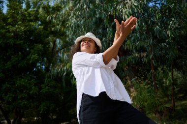 Neşeli bir kadın güneşli bir parkta beyaz gömlek ve şapkasıyla dans ediyor. Etrafı yeşilliklerle çevrili, mutluluk saçıyor..