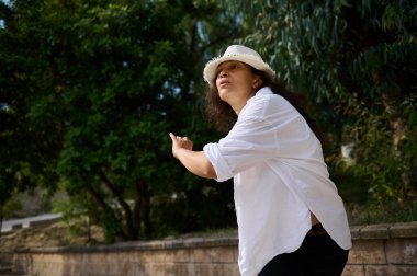 Beyaz gömlekli ve şapkalı bir kadın dışarıda dururken tutkulu bir jest yapıyor. Etrafı yemyeşil ağaçlarla çevrili. Huzurlu ve doğal bir ortam yaratıyor..