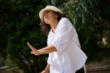 Beyaz şapka ve gömlek giymiş bir kadın dışarıda zarif bir şekilde dans ediyor. Yeşil arkaplan, onun etkileyici hareketlerini ve doğada olmanın neşesini vurguluyor..