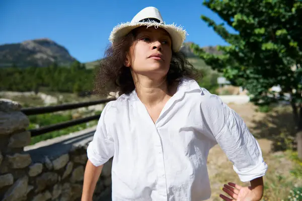 stock image Woman wearing a white shirt and hat, standing outdoors in a scenic mountain landscape. A sense of relaxation and freedom in nature on a sunny day.