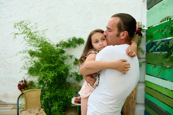 stock image A loving father holding and hugging his young daughter in a serene outdoor garden setting. The image conveys feelings of love, safety, and family bonding in a natural environment.