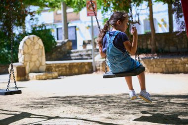 Kot pantolonlu, atkuyruklu, güneşli bir parkta neşeyle sallanan genç bir kız. Sakin dış mekan ağaçlar, bir taş yapı ve gölgeler içeriyor..