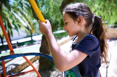 Neşeli bir genç kız, yüzünde bir gülümseme ile güneşli bir günde renkli bir oyun alanında oynuyor. Oyun ekipmanlarını elinde tutuyor ve dışarıda eğleniyor..
