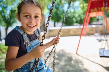 Güneşli bir parkta salıncakta gülümseyen neşeli genç kız kaygısız bir eğlence ve çocukluk mutluluğunu yakalıyor..