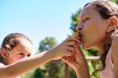 Parkta güneşli bir günde bir dondurma külahını paylaşan bir anne ve kızın sevgi, bağ ve neşe dolu bir anı..