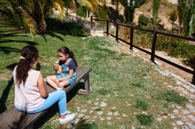 Anne ve kızı güzel bir parkta güneşli bir günde dondurma yiyerek bir neşe anlarını paylaşıyorlar. Açık havada aile bağı..