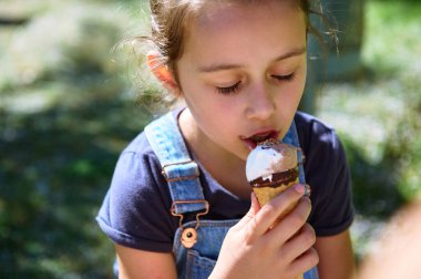 Açık havada dondurma külahının tadını çıkaran, parkta güneşli bir günün tadını çıkaran genç bir kızın yakın çekimi. Mavi bir gömlek ve mavi tulum giyiyor..