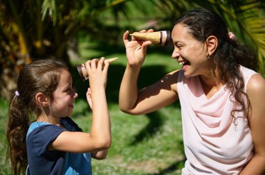 Anne ve kızının açık havada dondurma külahlarıyla oynayıp yeşil bir parkta güneşli bir günün tadını çıkardığı neşeli bir an. Aile bağı ve mutluluk.