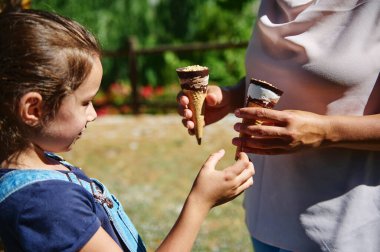Bir anne ve küçük kızı, güneşli bir parkta dondurma külahlarının keyfini çıkarırken, bir anlık bağ ve yaz eğlencesi sergiliyorlar..