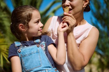 Bir anne ve kızı güneşli bir günün tadını çıkarıyor, dondurmayı paylaşıyor. Kız neşeli bir şekilde annesine bakıyor, sıcak ve neşeli bir an yaratıyor..