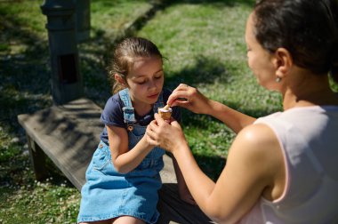 Bir anne ve kızı, parkta güneşli bir günün tadını çıkarırken, bir dondurma külahını paylaşır, ahşap bir bankta birlikte otururlar..