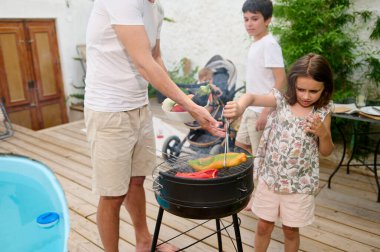 Aile, yaz gününün tadını çıkarıyor arka bahçede mangal yaparken sebze pişiriyor. Bir çocuk ızgaraya yardım ederken, diğeri bakar, beraberlik ve eğlence anını yakalar..