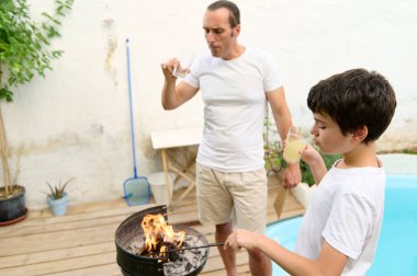 Baba ve oğul arka bahçede ızgara yapıyor, ikisi de ferahlatıcı limonatanın tadını çıkarıyor. Izgaranın etrafında bir yaz günü bağ kurma anı. Çocuk ateşe bakıyor..