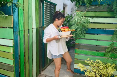 Elinde bir kase taze sebzeyle yeşil bir çitle canlı bir bahçede yürüyen bir kadın. Yaz günü, bahçe işleri ve sağlıklı yaşam konsepti..