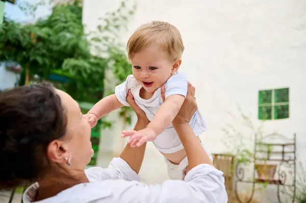 stock image Joyful mother lifting her smiling baby in the air, creating a loving moment in an outdoor setting with a white wall backdrop.