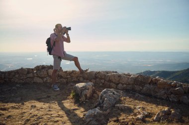 Dağın tepesinde kamerayla fotoğraf çeken bir adam. Günlük kıyafetler giyiyor, sırt çantası takıyor ve panoramik manzarayı seviyor..
