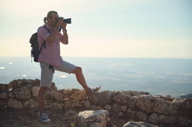 Sırt çantası ve kameralı bir seyahat fotoğrafçısı güneş doğarken bir dağın zirvesinden fotoğraf çekiyor ve manzarayı yakalıyor. Sahne, keşif, macera ve doğal güzelliği çağrıştırıyor..