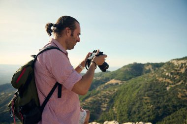 Sırt çantalı bir erkek fotoğrafçı, parlak bir günde, manzaralı bir dağ manzarasını incelerken kamerasını kontrol ediyor. Açık hava macerası ve doğa fotoğrafçılığı kavramları.
