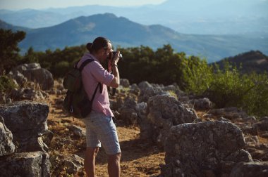 Sırt çantalı bir yürüyüşçü güzel bir dağ manzarasının fotoğrafını çeker. Görüntü macera ve açık hava keşiflerinin özünü yakalıyor kayalık arazi ve yemyeşil yemyeşil.