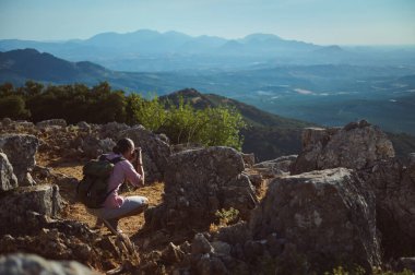 Bir fotoğrafçı gündüz vakti kayalık bir dağ manzarasında fotoğraf çeker. Açık mavi gökyüzü ve arka plandaki geniş sıradağlar çarpıcı bir doğa manzarası yaratıyor..